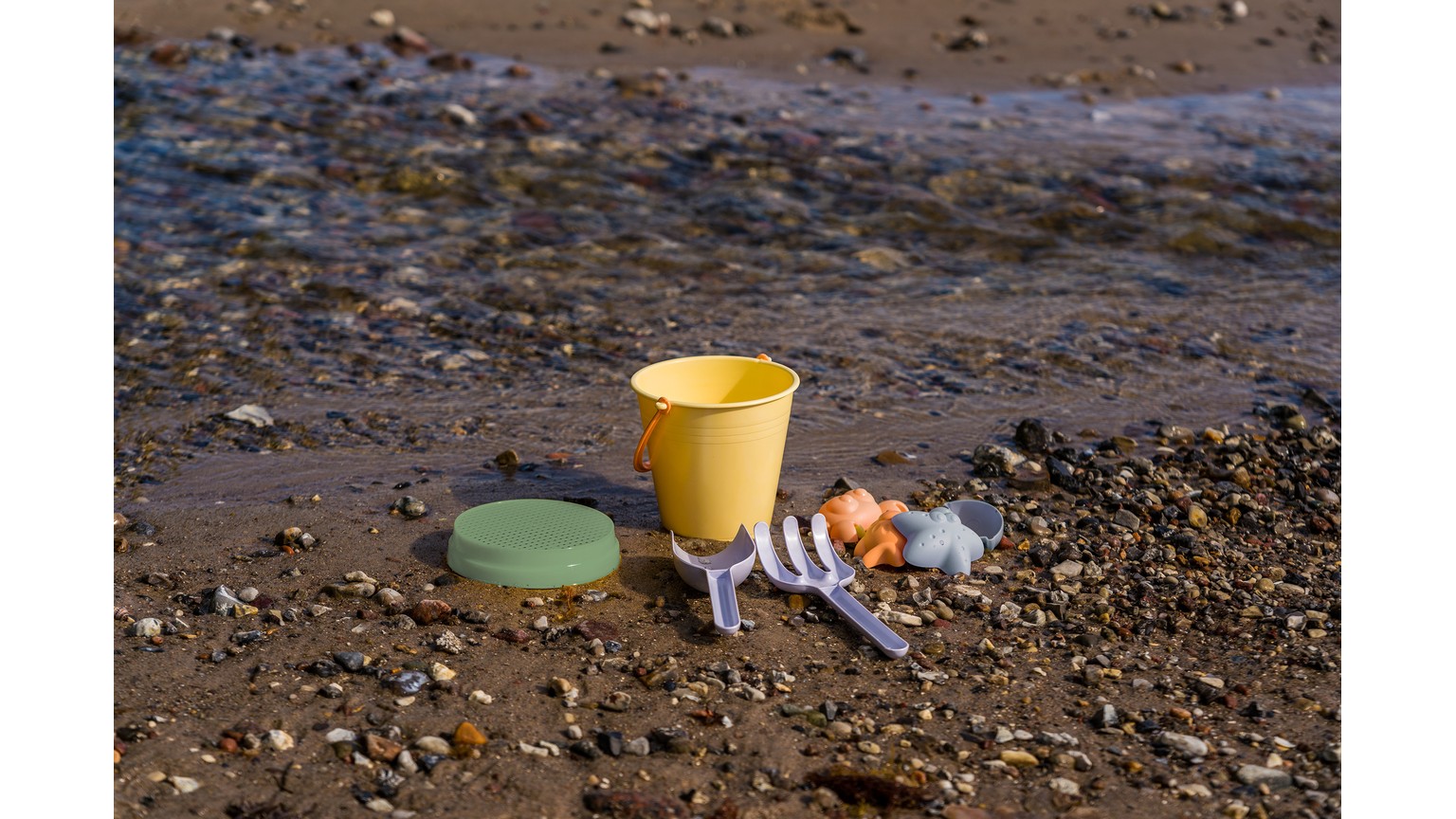 Gul spand og grøn skovl ligger på sandet ved vandkanten med små skaller omkring